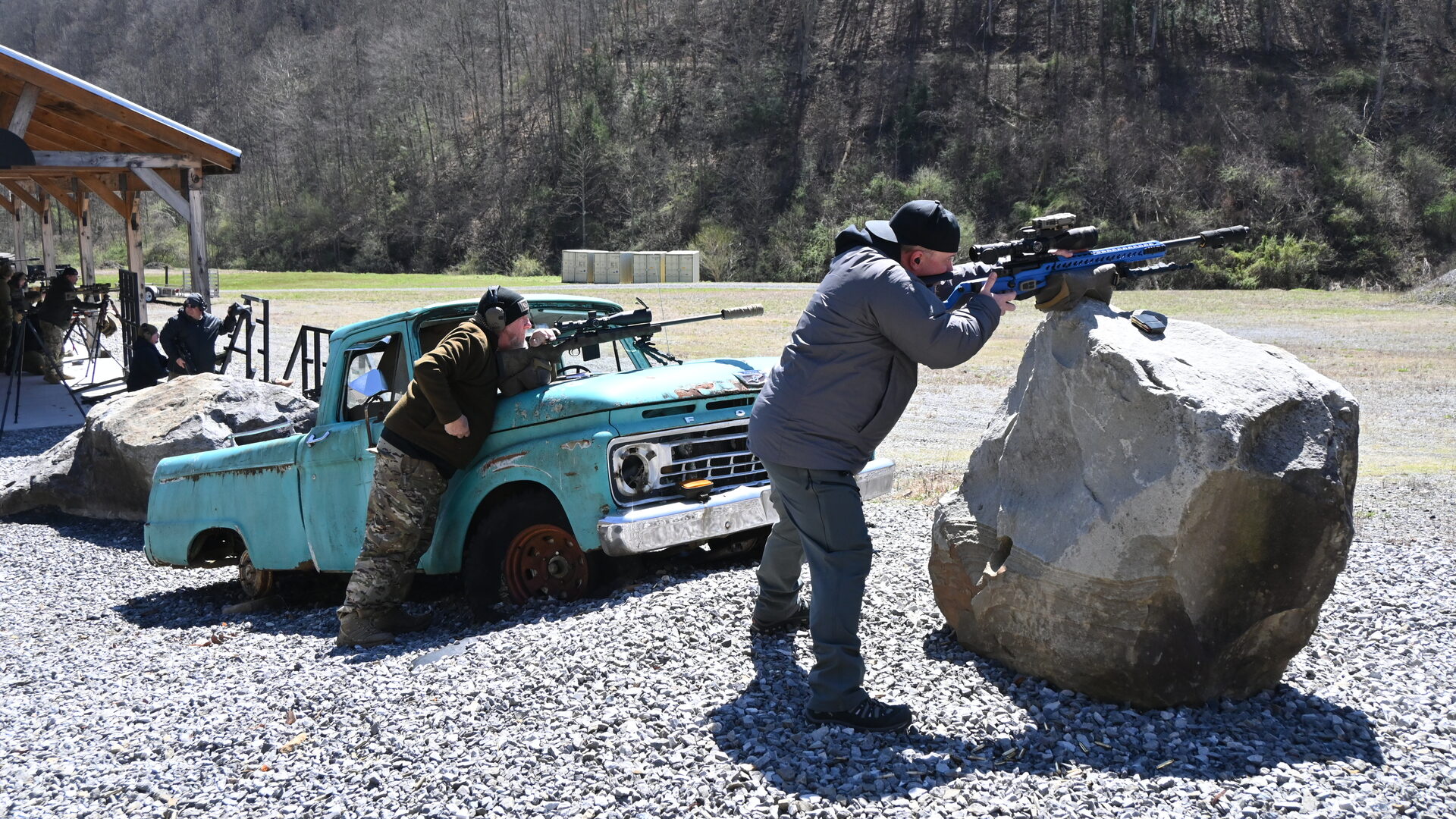 Mountain Rifleman 26 students working from improvised positions — one prone behind a turquoise pickup truck, the second standing braced against a boulder taking an offhand shot. Real obstacles. Real positions.